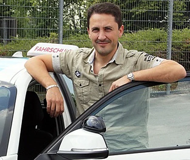 Der Inhaber der Fahrschule Geldi lehnt lächelnd an der Tür des Fahrschulautos